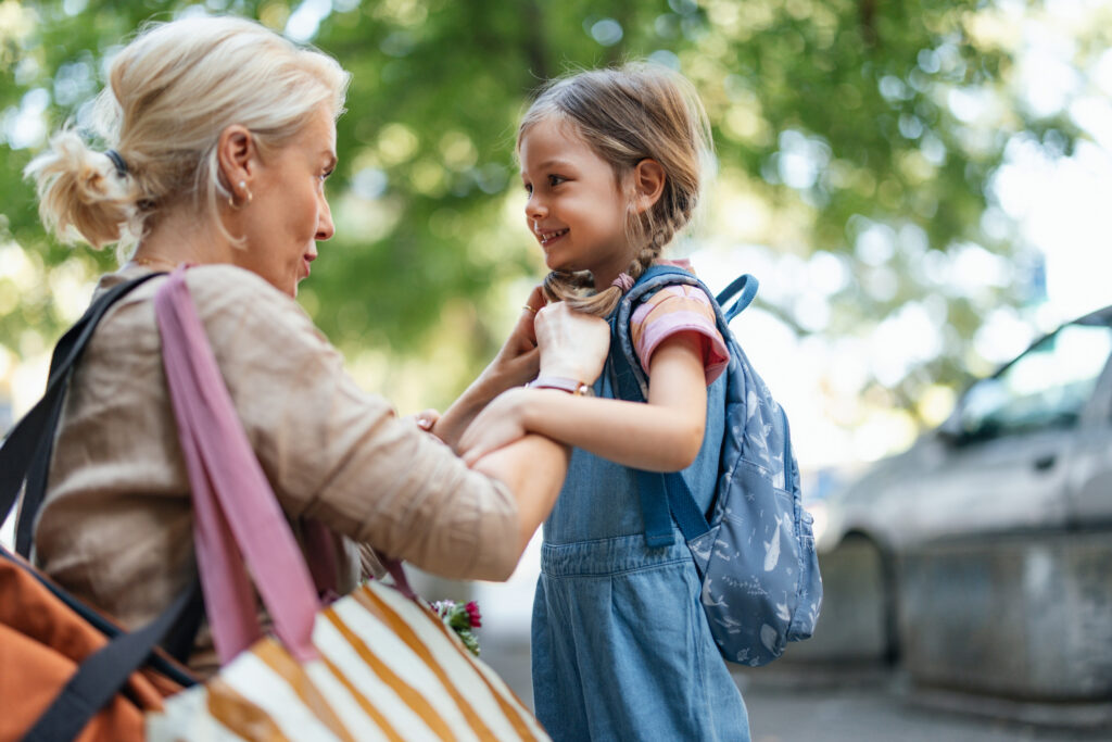 Woman crouching down to get young girl ready for school in an outdoor setting, both are smiling.